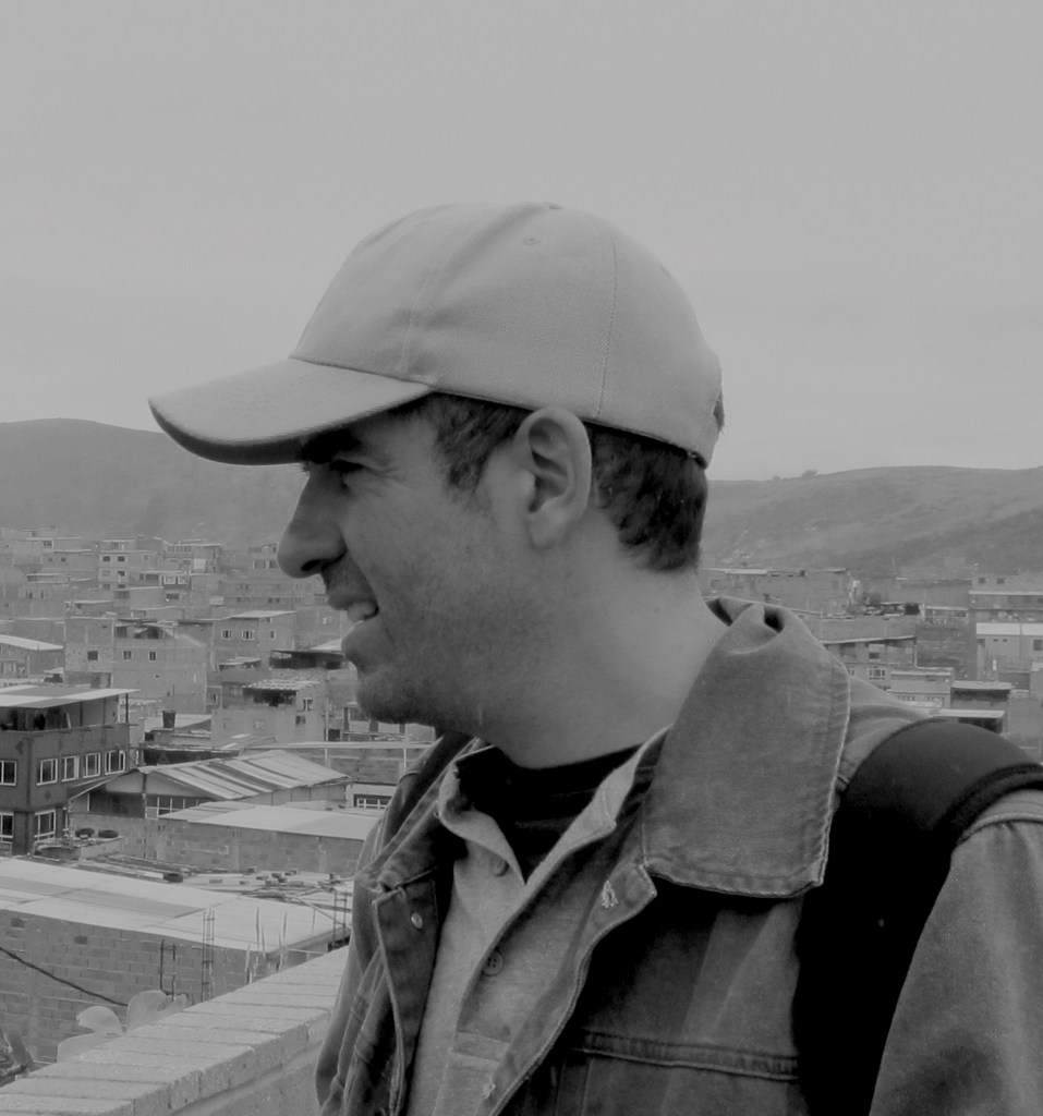 Black and white profile photo of white man with grey cap looking to the side. Behind a sprawling landscape of an urban periphery.